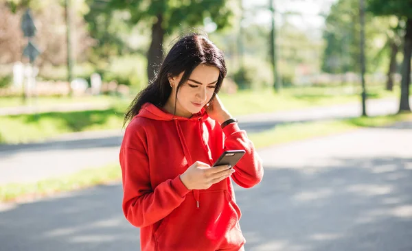 Stylish girl in red sweater posing on alley holding smartphone. Happy woman with beautiful smile chatting on cell smartphone while relaxing in the park, outdoors