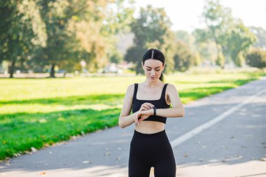 Siyah bluzlu şık bir kız. Saate bakıyor ve parkta dinleniyor. Açık havada, yaz zamanı. Sabahları koşan bir kadın. Açık hava antrenmanı. Spor ve sağlıklı aktif yaşam tarzı konsepti.