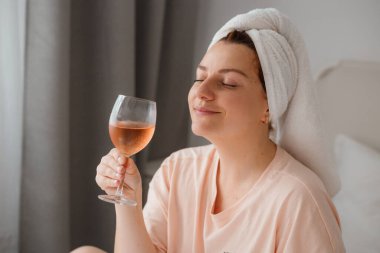 Duş aldıktan sonra yatak odasında bir bardak şarapla güzel, mutlu bir genç kadın. Karantina süresince evde tecrit altında kalmak. Doğum gününü yatakta yalnız kutlama kavramı