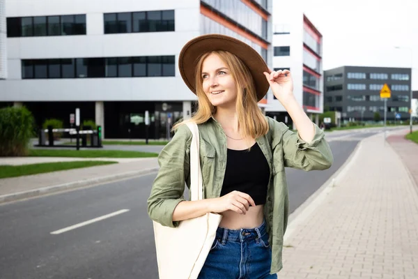 Portrét mladé šťastné ženy procházející se po ulici ve městě. Fotografie módní dívka s kloboukem a plátěnou taškou s úsměvem do kamery. — Stock fotografie