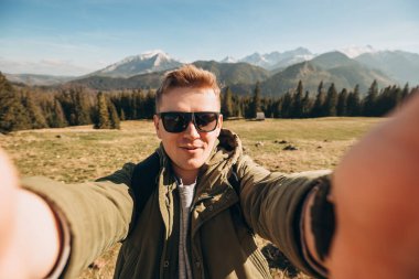 Güneş gözlüklü genç adam kar dağlarında fotoğraf çekiyor manzaranın tadını çıkarıyor. Özgürlük, mutluluk, seyahat ve tatil konsepti, açık hava aktiviteleri, yeşil ceket giyiyor.