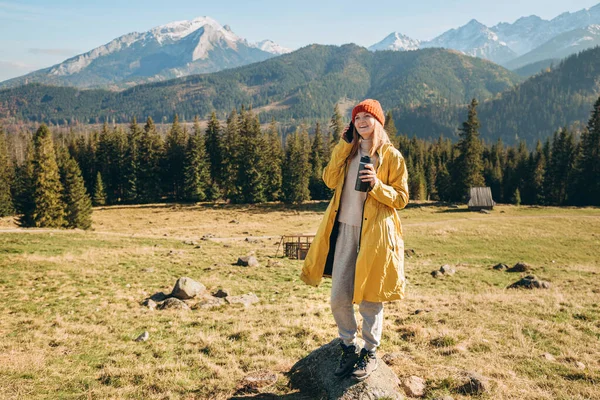 Νεαρή ξανθιά γυναίκα με θερμός μιλάει με smartphone στα βουνά. Ελευθερία, ευτυχία, ταξίδια και διακοπές έννοια, υπαίθριες δραστηριότητες. Ευτυχισμένο κορίτσι με κόκκινο καπέλο και κίτρινο μπουφάν. — Φωτογραφία Αρχείου