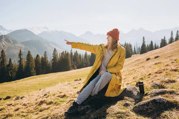 Joven turista apuntando a hermosas montañas y disfrutando de una vista increíble. Concepto de libertad, felicidad, viajes y vacaciones, actividades al aire libre, lleva sombrero rojo y chaqueta amarilla — Foto de Stock