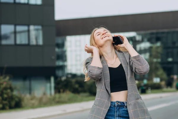 Portrét mladé šťastné blondýny s telefonem procházející se po ulici ve městě. Koncept technologie nebo lidí. Nadšený dívka se směje na bulding pozadí. — Stock fotografie