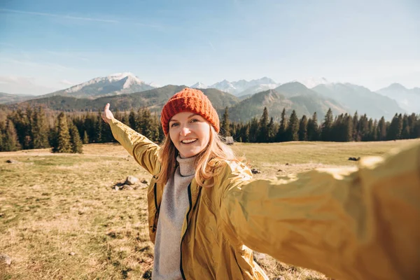 Νεαρή γυναίκα βγάζει selfie φωτογραφία ψηλά στα χιονισμένα βουνά απολαμβάνοντας τη θέα. Ελευθερία, ευτυχία, ταξίδια και διακοπές, υπαίθριες δραστηριότητες, φοράει κόκκινο καπέλο και κίτρινο μπουφάν. Ακολουθήστε με. — Φωτογραφία Αρχείου