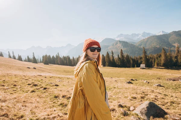 山の中でサングラスをかけて踊る若いブロンドの女性。自由、幸福、旅行や休暇の概念、屋外活動。ハッピーガールは赤い帽子と黄色のジャケットを着て — ストック写真