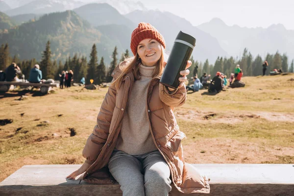 Young blond woman with thermos cup resting on the bench in mountains. Freedom, food, travel and vacations concept, outdoor activities. Happy Girl wearing red hat, halt — Stock Photo, Image