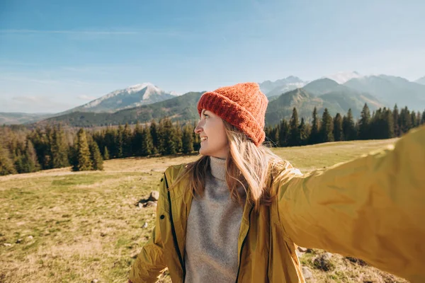 雪の山の中で自撮り写真を撮る若い女性が景色を楽しんでいます。自由、幸福、旅行や休暇のコンセプト、屋外活動、彼女は赤い帽子と黄色のジャケットを着ています — ストック写真