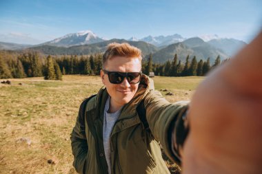 Güneş gözlüklü genç adam kar dağlarında fotoğraf çekiyor manzaranın tadını çıkarıyor. Özgürlük, mutluluk, seyahat ve tatil konsepti, açık hava aktiviteleri, yeşil ceket giyiyor.