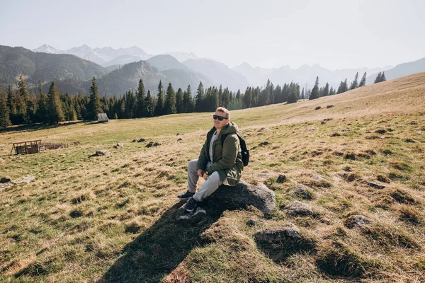 Young man in sunglasses with backpack see view on top mountain. Freedom, happiness, travel and vacations concept, outdoor activities. — Stock Photo, Image