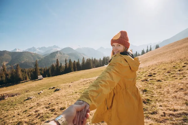 Hiker with red hat stands on edge of cliff against background and extends a hand. Happy woman gives a hand to someone like follow me. First person view. — Stock Photo, Image