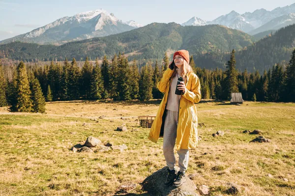 Mujer rubia joven con termo taza hablando por teléfono inteligente en las montañas. Concepto de libertad, felicidad, viajes y vacaciones, actividades al aire libre. Chica feliz con sombrero rojo y una chaqueta amarilla — Foto de Stock