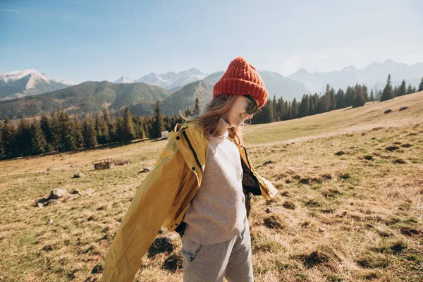 Young blond woman with sunglasses dancing in mountains. Freedom, happiness, travel and vacations concept, outdoor activities. Happy Girl wearing red hat and a yellow jacket. Wind in the hair. — Stock Photo, Image