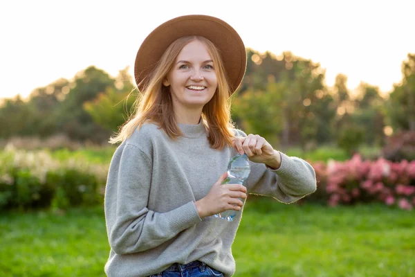 Primer plano retrato de una atractiva joven rubia con sombrero bebiendo agua de botella de plástico en el parque verde de verano. Concepto de alimentación, descanso o ecología — Foto de Stock