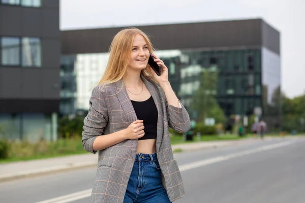 Portrét mladé šťastné blondýny s telefonem procházející se po ulici ve městě. Koncept technologie nebo lidí. Nadšený dívka usmívá na bulding pozadí. — Stock fotografie
