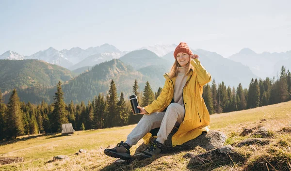 Traveler girl drinking tea from thermos cup over nature background. Mountain peaks. Freedom, happiness, travel and vacations concept, outdoor activities, woman wearing red hat — Stock Photo, Image