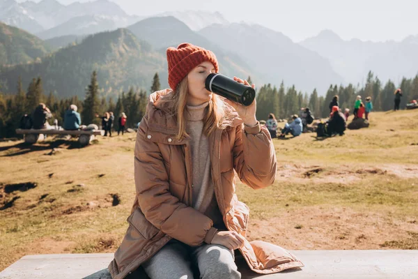 Μια νεαρή ξανθιά γυναίκα με θερμός που αναπαύεται στον πάγκο στα βουνά. Ελευθερία, φαγητό, ταξίδια και διακοπές έννοια, υπαίθριες δραστηριότητες. Ευτυχισμένο κορίτσι που φοράει κόκκινο καπέλο, αλτ — Φωτογραφία Αρχείου