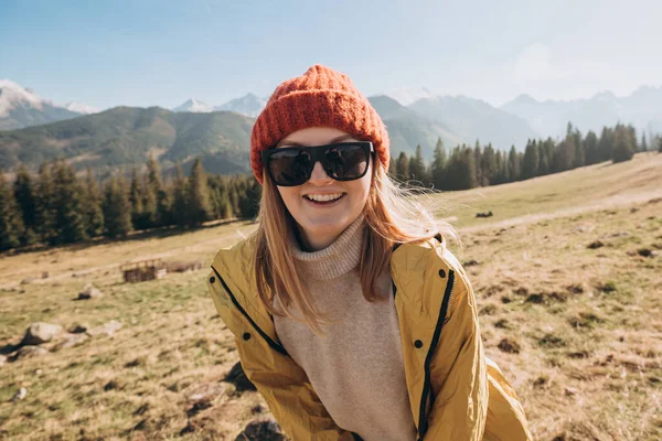 Fiatal szőke nő napszemüvegben, aki a hegyekben táncol. Szabadság, boldogság, utazás és nyaralás koncepció, szabadtéri tevékenységek. Boldog lány piros kalapban és sárga kabátban. — Stock Fotó