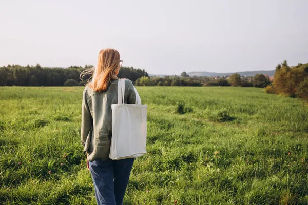 Mujer rubia joven con bolsa de algodón blanco en las manos sobre fondo natural. Bolsa de lona reutilizable vacía mockup para el diseño. — Foto de Stock