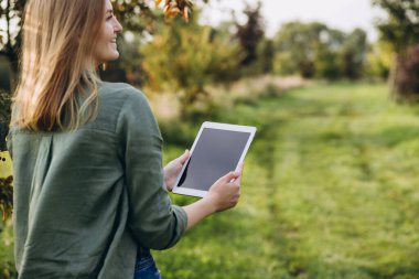 Elinde tablet olan bir çiftçi yeşil bahçede duruyor. Tablet ekranına bakıp gülümseyen bir kadın. Modern yaşam tarzı ve rahatlama kavramları. Model
