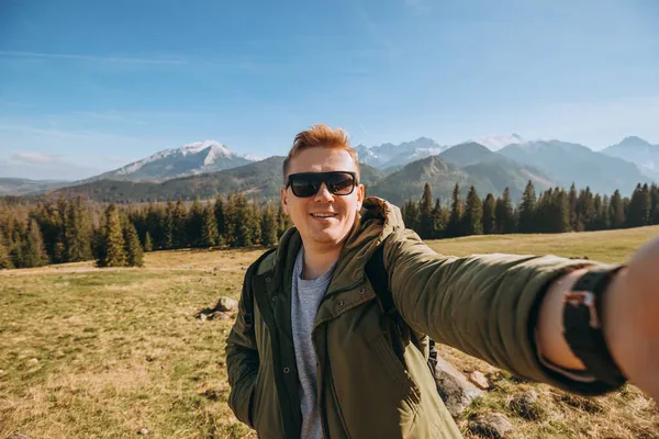 雪の山の中で自撮り写真を撮る若い男性が景色を楽しんでいます。自由、幸福、旅行や休暇の概念、屋外活動、彼は緑のジャケットを着て — ストック写真
