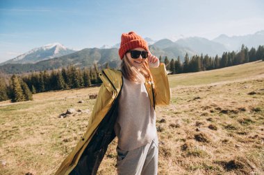 Dağlarda dans eden güneş gözlüklü genç sarışın kadın. Özgürlük, mutluluk, seyahat ve tatil konsepti, açık hava aktiviteleri. Kırmızı şapkalı ve sarı ceketli mutlu kız.