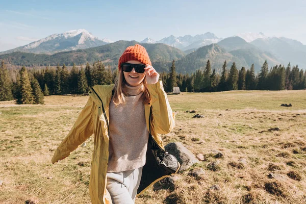 Dağlarda dans eden güneş gözlüklü genç sarışın kadın. Özgürlük, mutluluk, seyahat ve tatil konsepti, açık hava aktiviteleri. Kırmızı şapkalı ve sarı ceketli mutlu kız. — Stok fotoğraf