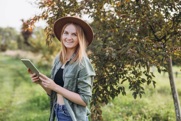 Farmářka s tabulkou v ruce stojí v zelené zahradě. Žena se usmívá a dívá se do kamery. Moderní životní styl a relaxační koncepce. — Stock fotografie