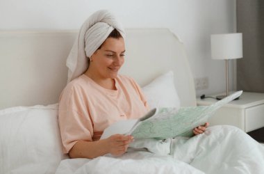 Yatağında haritası olan mutlu genç bir kadın. Olumlu gülümseyen kız evde ya da otelde oturur. Turist seyahati ve rahatlama konsepti.