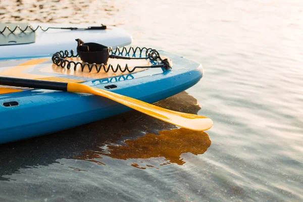 Placa de remo amarela e duas pranchas de surf em fundo azul superfície de água limpa. Surf e SUP equipamento de embarque em luzes de pôr do sol close-up. Esportes aquáticos ao ar livre. Conceito de estilo de vida surf. — Fotografia de Stock