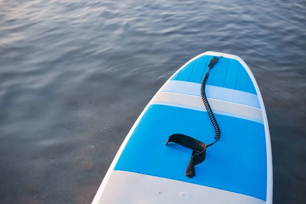 Deska surfingowa na niebieskim tle czystej wody. SUP wyposażenie pokładowe w świetle zachodzącego słońca zbliżenie. Sporty wodne na świeżym powietrzu. Wynajem sprzętu do pływania w oceanie lub morzu, czas letni — Zdjęcie stockowe