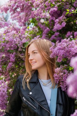Sarı saçlı, mavi bluzlu ve siyah ceketli genç bir kadın bahar parkında yürüyor. Leylak kokusunun tadını çıkaran güzel bir kadın. Olumlu duygular ve hisler. Tatil konsepti, Kadınlar Günü pankartı