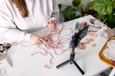 Kadın oturma odasında yeni video bloğu kaydetti. Akıllı telefonu tripodlu kıza takıp çevrimiçi canlı yayın yapıyor. Webinar 'dan uzak sohbetler, modern teknoloji, video konsepti. İş sırasında el yapımı makrome. Kadın hobisi