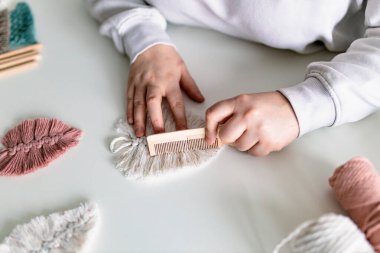 Neredeyse bitmiş bir makrome parçasının son detaylarını hazırlayan bir kadın, evin içinde ipleri tarıyor. Kapatın. Doğal pamuk iplikler ve tahta boncuklar. Kadın hobisi. Duvarda asılı dekor.