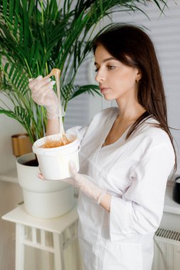 Eldivenli gülen epilasyon ustası plastik kavanozdaki şeker macununu esnetiyor. Balmumu epilasyon spa prosedürü. Kaplıca salonundaki ahşap bir çubukta tüy dökmek için sıvı sarı şeker veya balmumu.