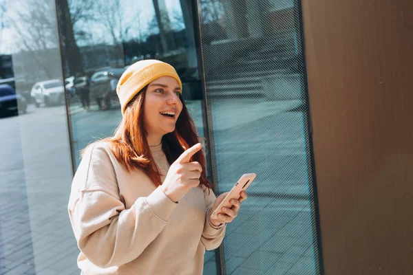 Şehir caddesinde yürüyen neşeli genç kadın akıllı telefonunu kontrol ediyor. Şehir yaşam tarzı konsepti. Gezgin. Güzel moda kızı dışarıda yürümeyi işaret ediyor
