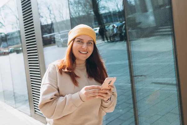 Şehir caddesinde yürüyen neşeli genç kadın akıllı telefonunu kontrol ediyor. Açık havada akıllı telefon kullanan sarı şapkalı güzel bir kadının portresi. Şehir yaşam tarzı konsepti. Gezgin