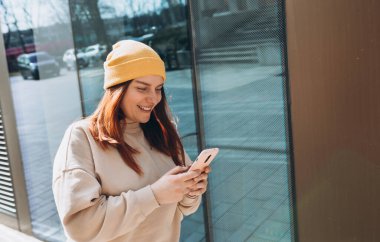 Şehir caddesinde yürüyen neşeli genç kadın akıllı telefonunu kontrol ediyor. Açık havada akıllı telefon kullanan sarı şapkalı güzel bir kadının portresi. Şehir yaşam tarzı konsepti. Gezgin
