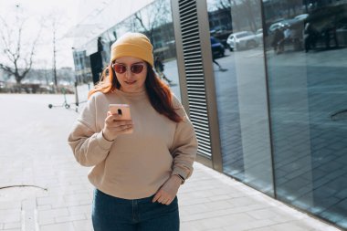 Şehir caddesinde yürüyen neşeli genç kadın akıllı telefonunu kontrol ediyor. Açık havada akıllı telefon kullanan, pembe güneş gözlüklü, sarı şapkalı güzel bir kadının portresi. Şehir yaşam tarzı konsepti. Gezgin