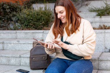 Açık havada tablet kullanan güzel, gülümseyen bir kız öğrenci. Şehir caddesinde merdivenlerde oturan neşeli genç bir kadın. Şehir yaşam tarzı konsepti. Gezgin, Çevrimiçi Eğitim veya Alışveriş