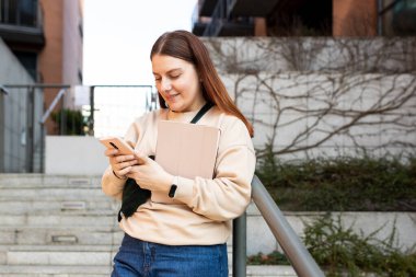 Şehir caddesinde yürüyen tabletli mutlu genç kadın akıllı telefonunu kontrol ediyor. Akıllı telefon kullanan güzel bir kadının portresi. Şehir yaşam tarzı konsepti. Gezgin. Çevrimiçi eğitim veya alışveriş