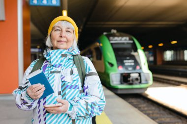 Sırt çantalı, sarı şapkalı yaşlı mutlu kadın biletini kontrol ediyor ve arka plandaki trende akıllı telefon kullanıyor. Demiryolu taşımacılığı konsepti. Demiryolu taşımacılığı kavramı