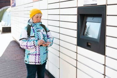 Yaşlı kadın paketi paket dolabından almış olmalı. Gülümseyen kadın, şehir caddesinde otomatik posta terminalinin yanında akıllı telefon tutuyor. Akıllı hızlı teslimat kavramı.