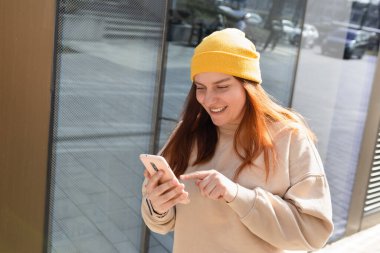 Şehir caddesinde yürüyen neşeli genç kadın akıllı telefonunu kontrol ediyor. Açık havada akıllı telefon kullanan sarı şapkalı güzel bir kadının portresi. Şehir yaşam tarzı konsepti. Gezgin