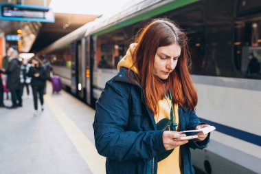 Sırt çantalı genç kızıl saçlı kadın biletini kontrol ediyor ve arka plandaki trende akıllı telefon kullanıyor. Demiryolu taşımacılığı konsepti. Bulanık tren istasyonu olan bir platformun yanında duran kız..