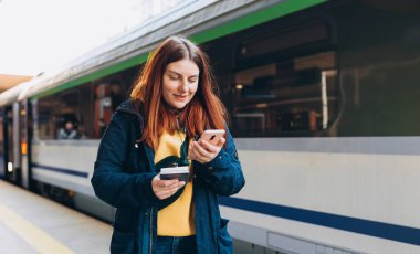Genç kızıl saçlı kadın arka plandaki elektrikli trende akıllı telefon kullanarak istasyon platformunda bekliyor. Demiryolu taşımacılığı kavramı
