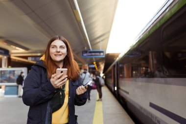 Genç kızıl saçlı kadın arka plandaki elektrikli trende akıllı telefon kullanarak istasyon platformunda bekliyor. Demiryolu taşımacılığı kavramı