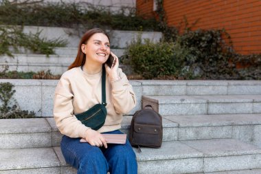 Güzel gülümseyen bayan öğrenci dışarıda telefonla konuşuyor. Telefon iletişimi. Şehir caddesinde yürüyen neşeli genç bir kadın, şehir yaşam tarzı konsepti. Gezgin