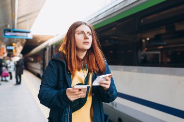 Genç kızıl saçlı kadın arka plandaki elektrikli trende akıllı telefon kullanarak istasyon platformunda bekliyor. Demiryolu taşımacılığı konsepti, Gezgin