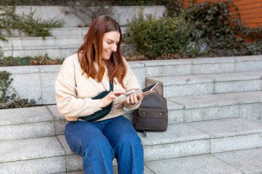 Açık havada tablet kullanan güzel, gülümseyen bir kız öğrenci. Şehir caddesinde merdivenlerde oturan neşeli genç bir kadın. Şehir yaşam tarzı konsepti. Gezgin, Çevrimiçi Eğitim veya Alışveriş